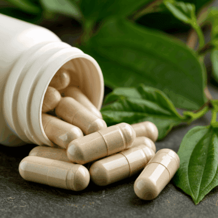 Open white bottle spilling beige capsules onto a dark surface with green leaves in the background suggesting dietary supplements