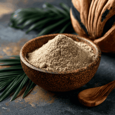 Beige powder in a wooden bowl on a dark surface with a wooden scoop and green palm leaves in the background